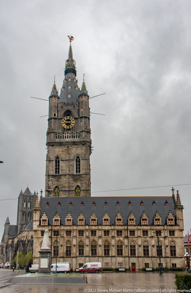 Ghent Belfry with clock tower and spire rising behind the long Gothic Cloth Hall under an overcast sky.