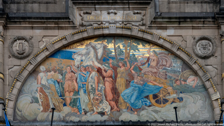 Large arched mural crowded with historical and allegorical figures, banners, animals, and a chariot.