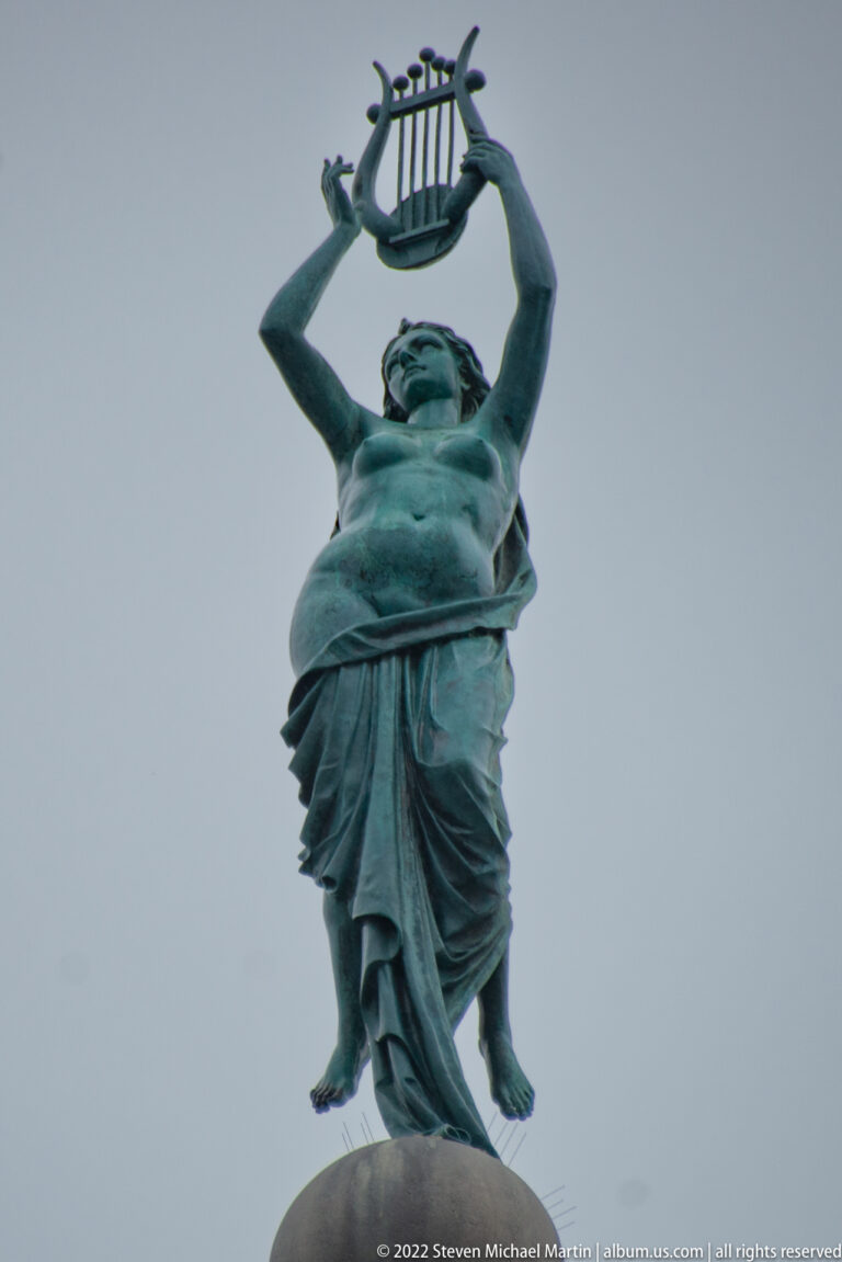 Bronze female statue in flowing drapery holding a lyre above her head against a gray sky.