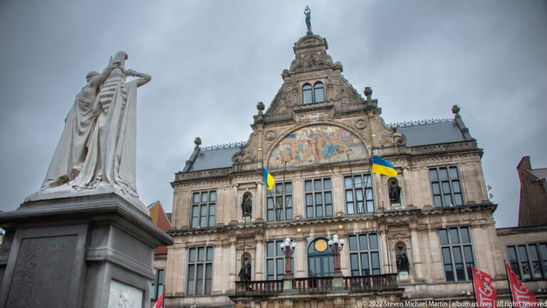 Ornate historic theatre façade with mural pediment, balcony flags, tall windows, and sculpture at the edge of the square.