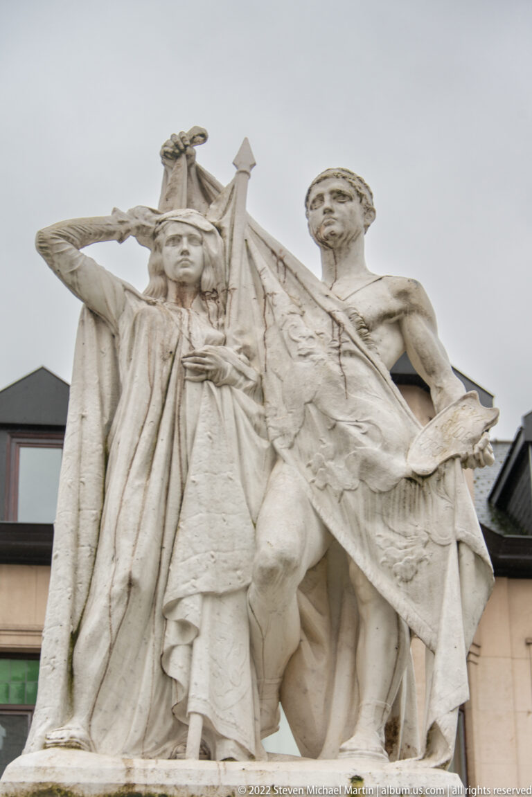 Large pale stone sculptural group of two classical figures with sweeping drapery viewed from below.
