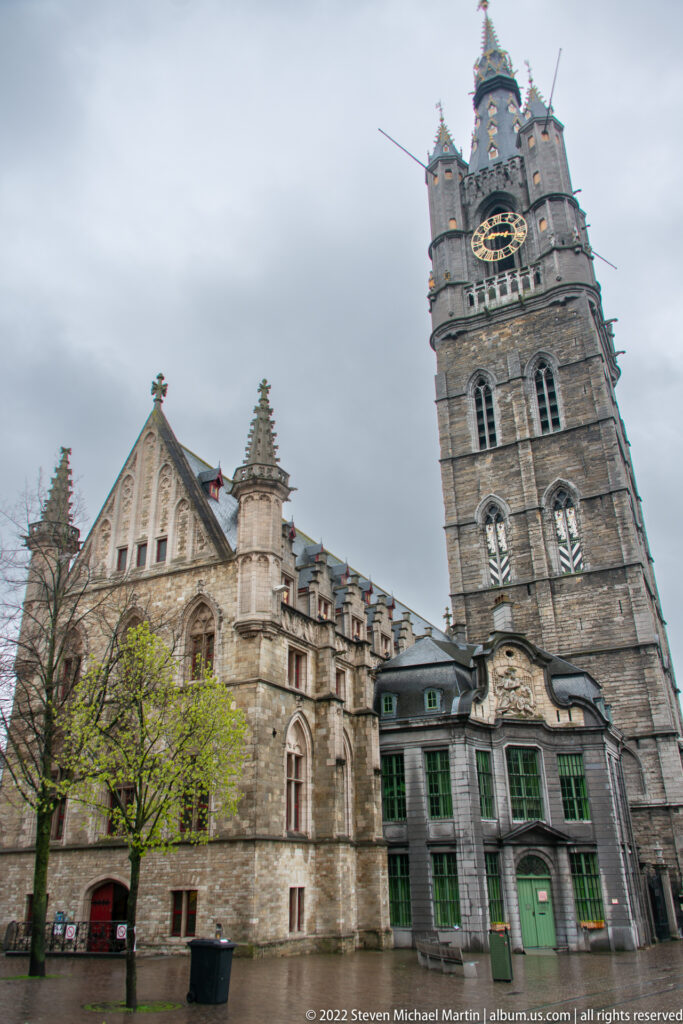 Ghent Belfry with clock and turrets rising beside lower Gothic stone buildings and a leafy tree.