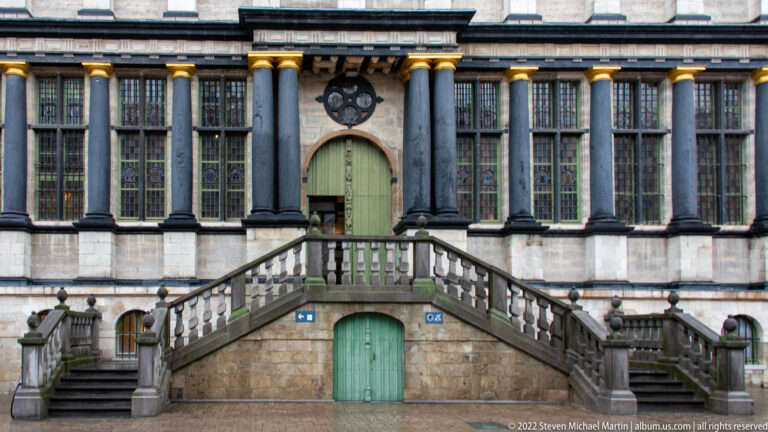 Symmetrical stone staircase leading to a pale green door between tall dark columns and large historic windows.