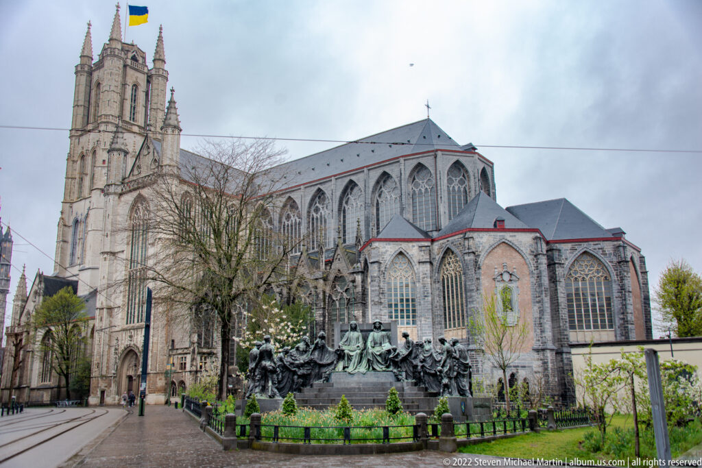Saint Bavo's Cathedral in Ghent with its tall west tower, Gothic windows, and a monument in the foreground garden.