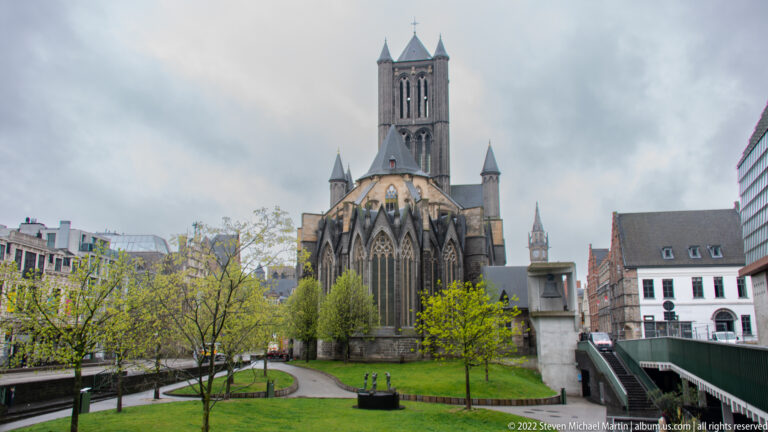 Saint Nicholas' Church in Ghent with central tower, Gothic windows, buttressed east end, trees, and landscaped foreground.