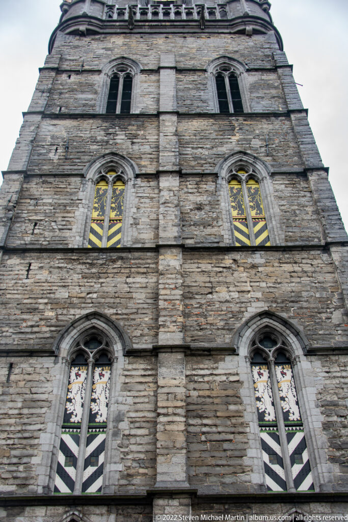 Upward close view of Ghent Belfry showing weathered stone courses, pointed windows, and colored glazing.
