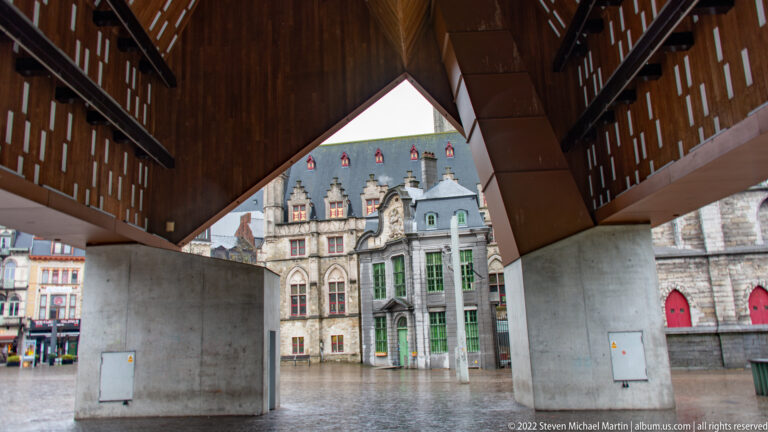 View beneath Ghent's Stadshal with timber roof and concrete piers framing ornate historic façades across the square.