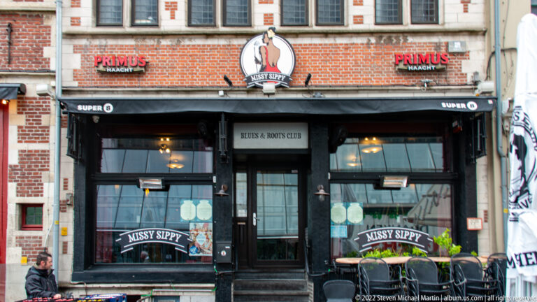 Black café storefront in a red-brick building with large windows, beer signs, and outdoor chairs.