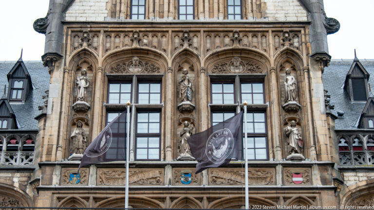 Close view of former Ghent post office windows framed by statues, tracery, shields, and stone carving with flags in front.