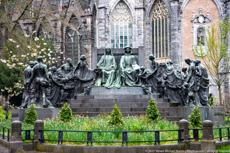 Large bronze multi-figure monument in a flowered garden with Gothic cathedral windows behind it.
