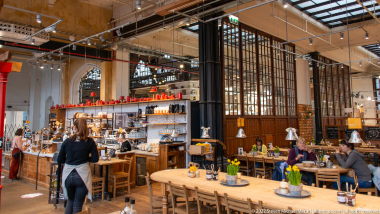 Busy indoor food hall with diners, wooden tables, retail counters, tall iron partitions, and an exposed ceiling.