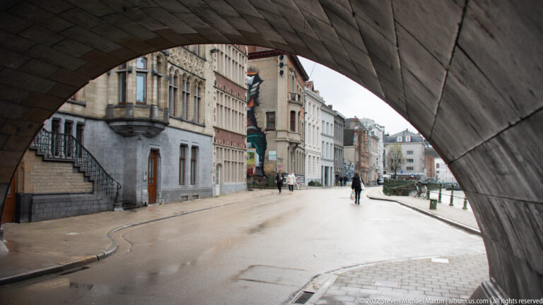 Historic Ghent street with pedestrians and façades framed by the underside of a large stone arch.