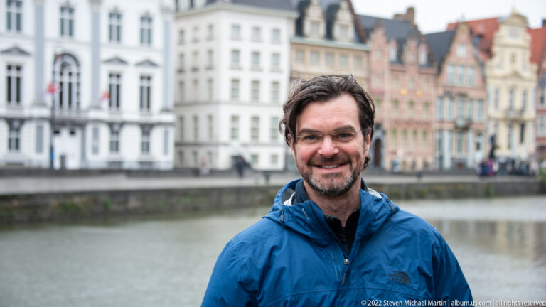 Smiling man in a blue jacket beside a Ghent canal, with softly blurred historic façades across the water.
