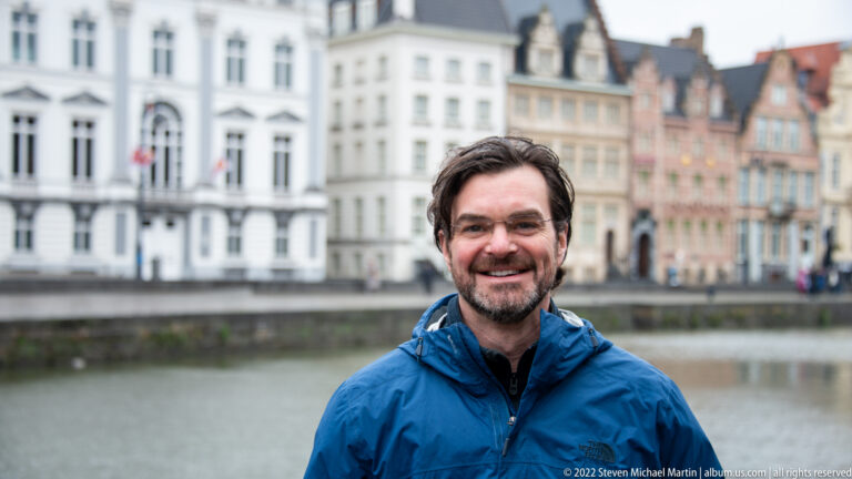Close portrait of a smiling man in a blue jacket with a Ghent canal and historic houses blurred behind.
