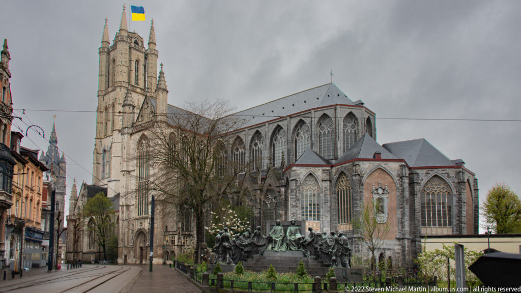 Wide street view of Saint Bavo's Cathedral showing the west tower, long Gothic nave, and monument beside the church.