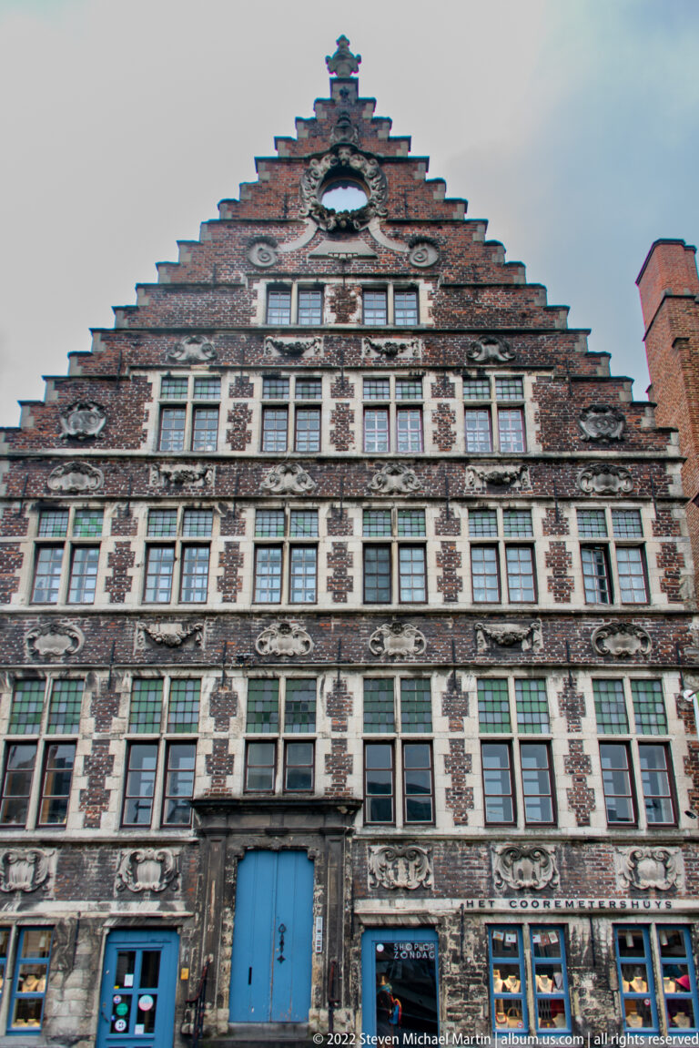 Large stepped-gable historic façade of dark brick and pale stone with rows of windows and blue doors.