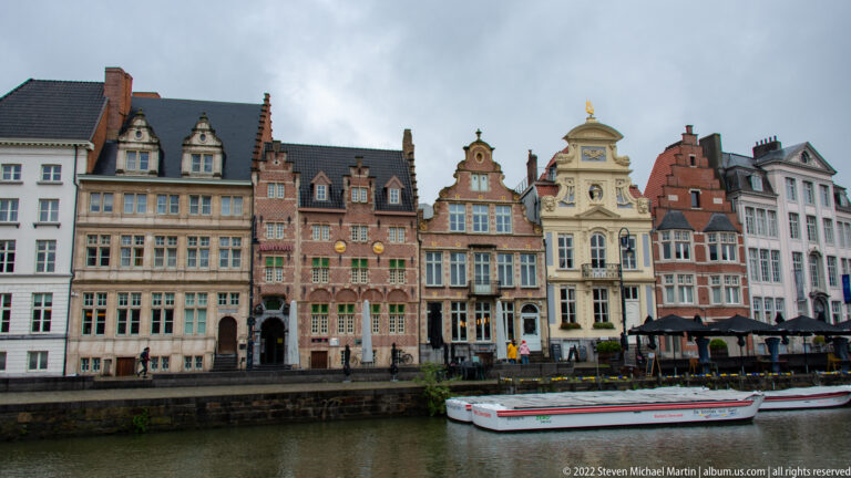 Colorful historic gabled houses lining a Ghent canal with a low passenger boat in front.