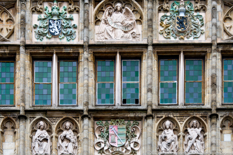Leaded windows framed by carved stone figures, heraldic shields, foliage, and relief panels.
