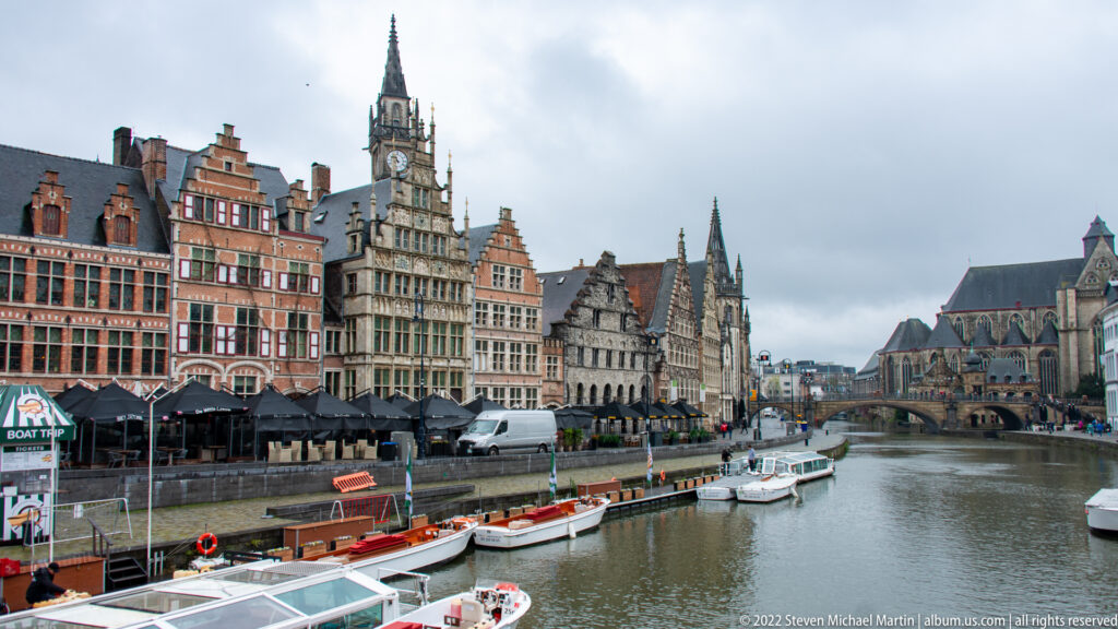 Wide Ghent canal view with moored boats, gabled waterfront houses, bridges, church roofs, and a tall central tower.