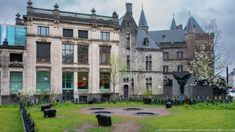 Historic stone building with pointed turrets beside a landscaped court, benches, lawn, and public sculpture.