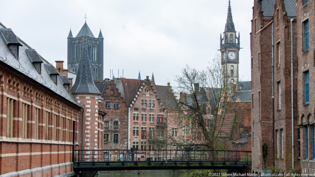 Historic Ghent skyline with multiple towers rising above brick buildings and a narrow bridge in the foreground.