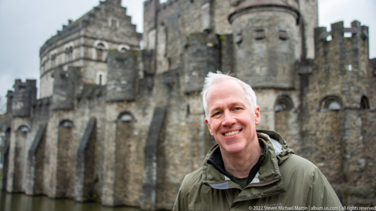 Smiling man in a green jacket standing before the softly blurred stone towers of Gravensteen castle.
