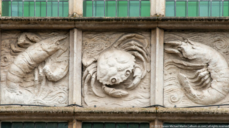 Weathered stone relief with a central grotesque face surrounded by swirling carved forms beneath a window band.