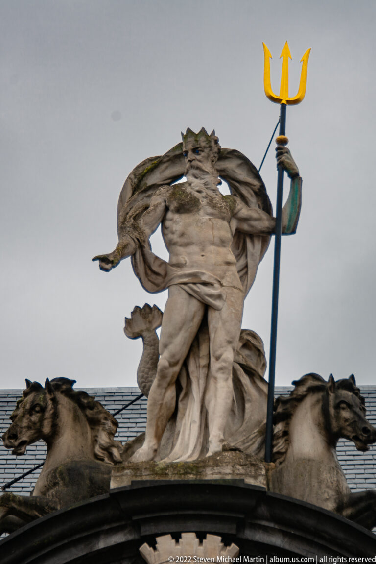 Stone Neptune figure holding a golden trident above two large sea creatures against a gray sky.