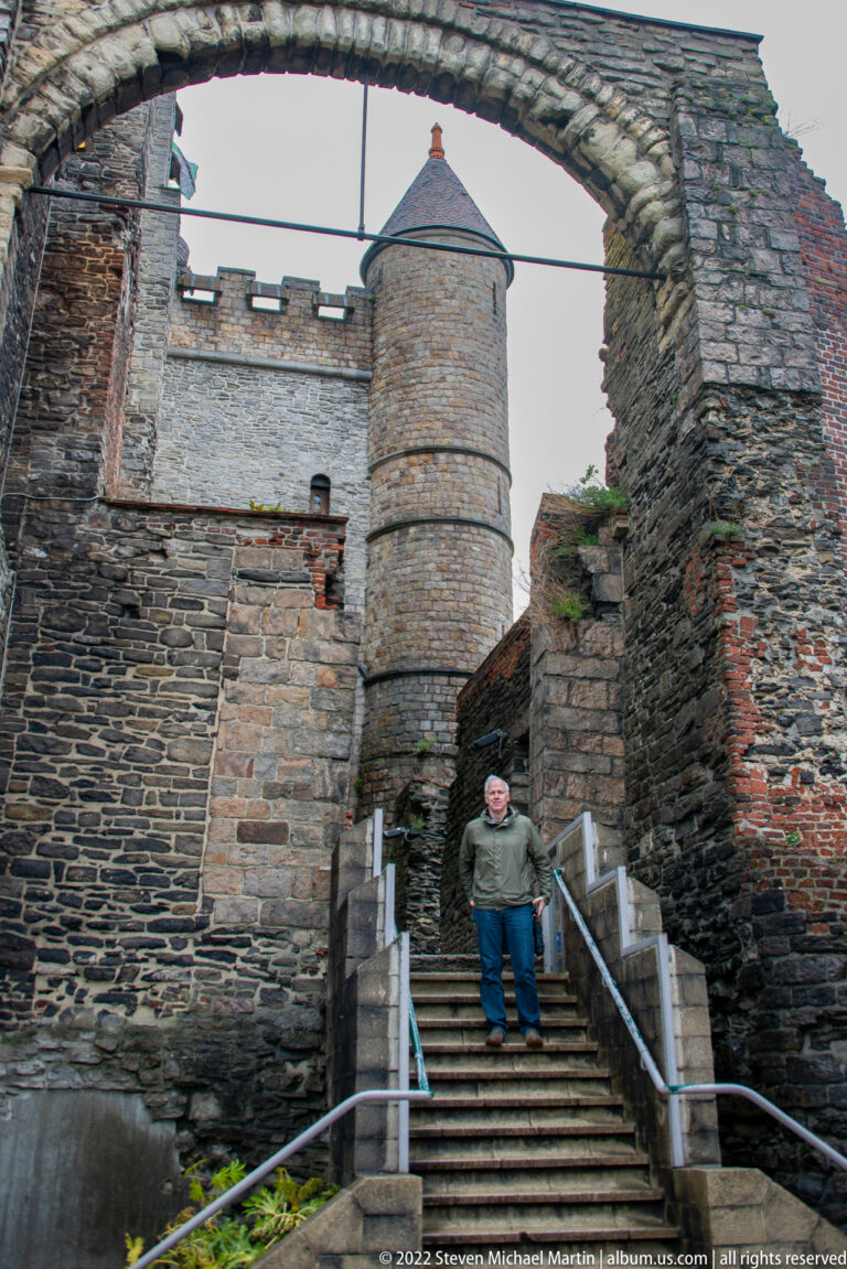 Stone stairs inside Gravensteen leading toward a round tower beneath a rough masonry arch, with one seated visitor.