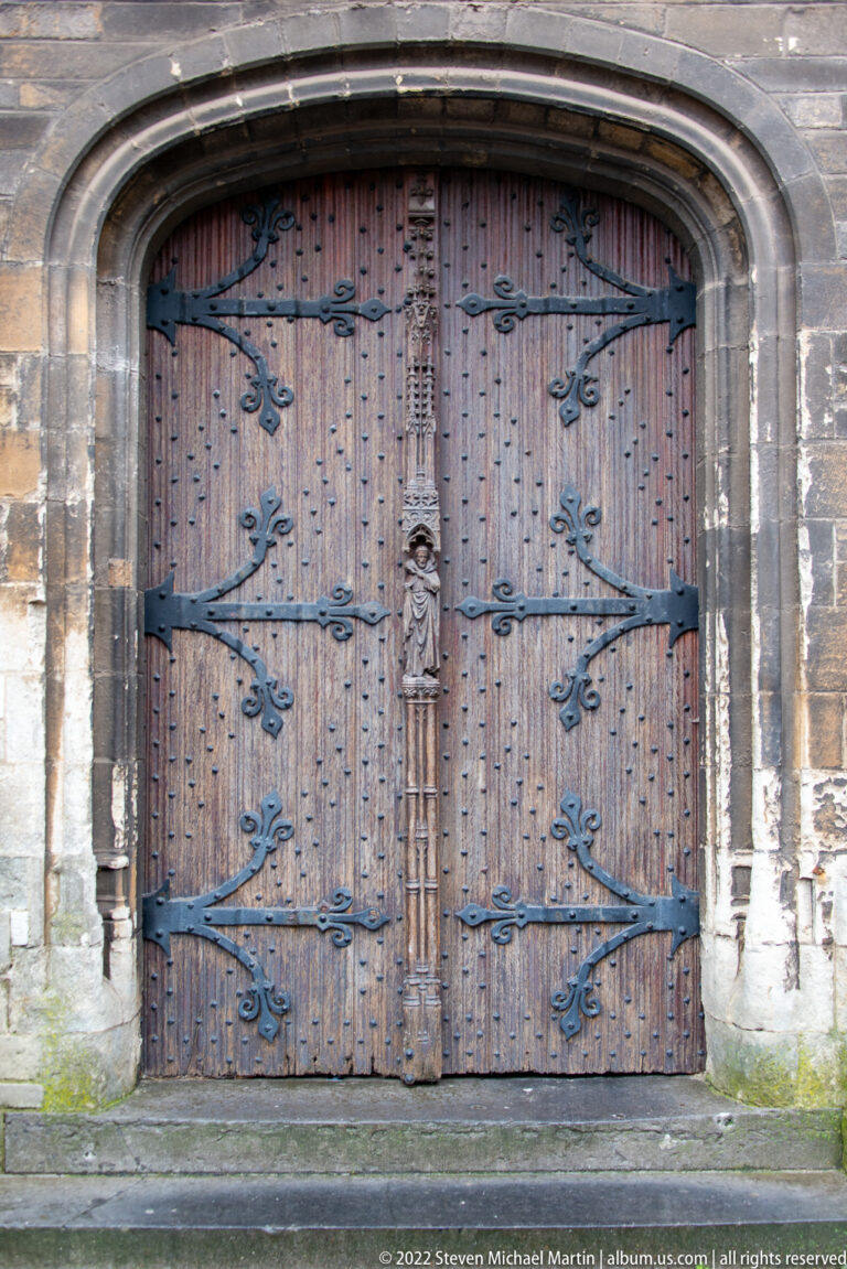Large arched wooden church door covered with metal studs and ornate wrought-iron strap hinges.