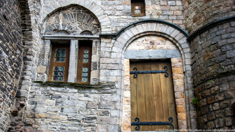 Timber door with black iron hinges beneath a stone arch beside a narrow leaded window inside Gravensteen.