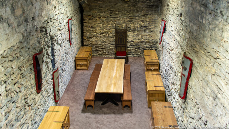 Long wooden table and benches in a narrow stone chamber with rough walls, small wall displays, and a dark timber door.