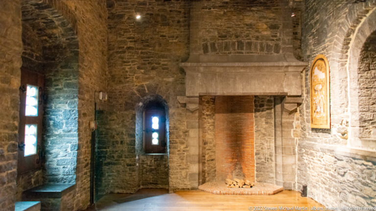 Large stone fireplace in a rough-walled castle room, flanked by narrow windows and lit warmly from the right.