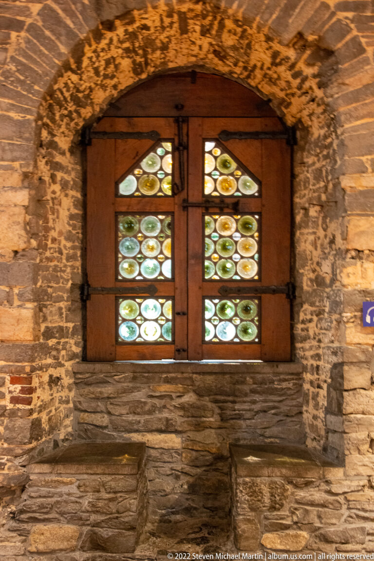 Small wooden-framed window filled with circular colored glass panes set deep in a rough stone castle wall.