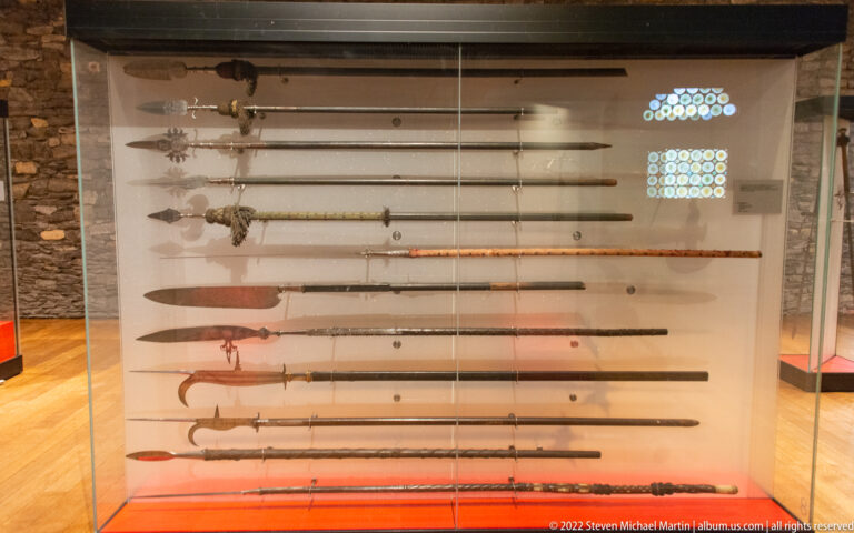 Glass case displaying rows of long spears, halberds, and other pole weapons mounted horizontally.