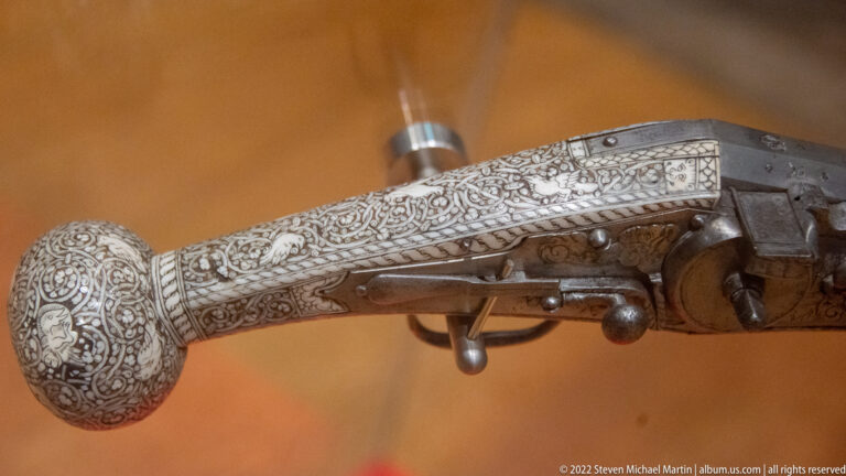 Close view of an ornate historic firearm with dense pale inlay, engraved metalwork, lock mechanism, and trigger.