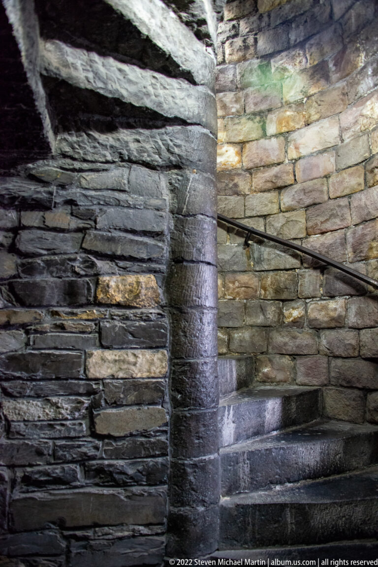 Narrow stone spiral staircase curving upward between rough masonry walls with a simple metal handrail.