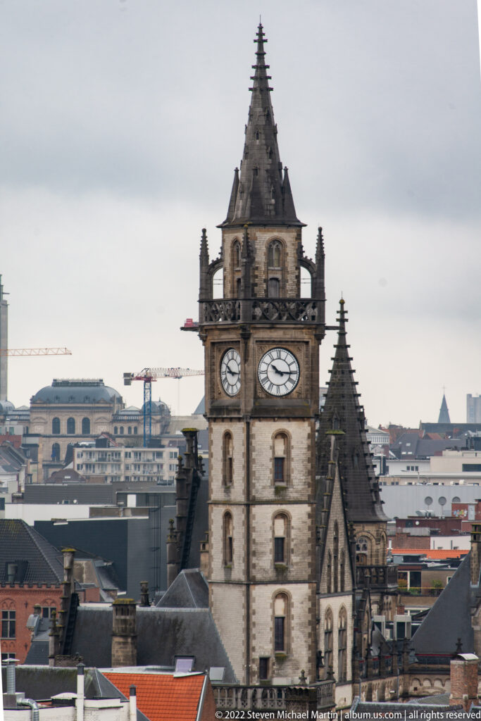 Historic Ghent tower with large clock faces and a steep tiered roof rising above city rooftops.