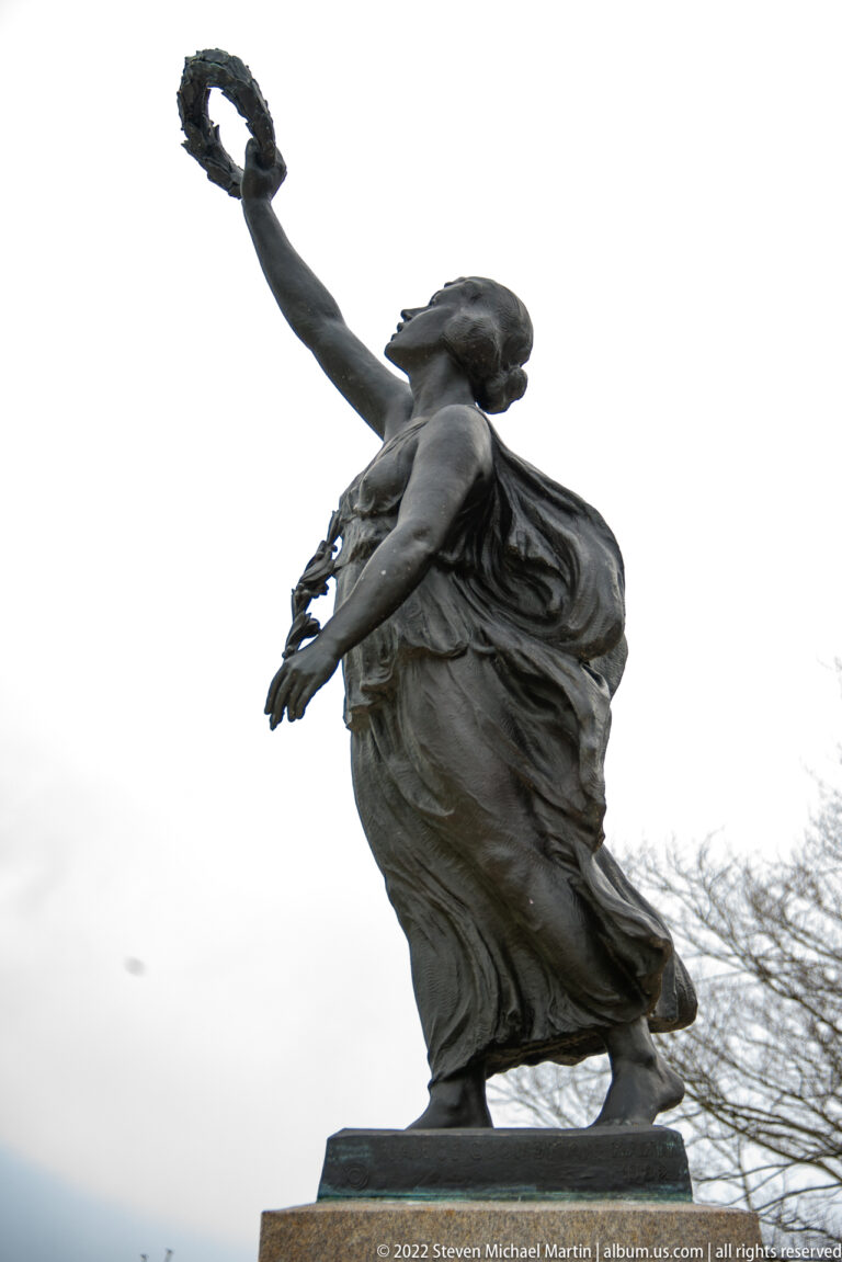 Bronze female allegorical figure leans forward with one arm raised and flowing drapery against the sky.