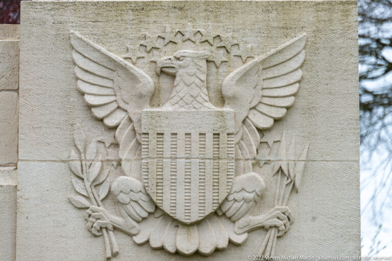 Carved American eagle with shield, arrows, olive branch, and stars on a pale stone wall.