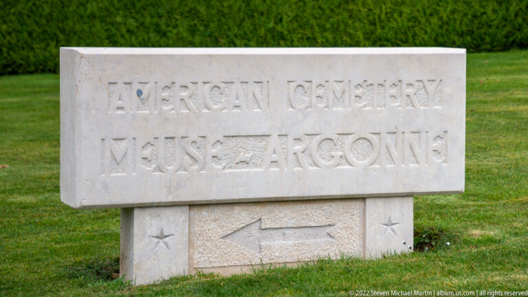 Stone sign reading American Cemetery Meuse-Argonne with a directional arrow, set on a green lawn.