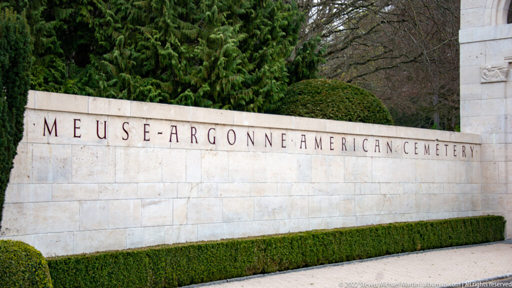 Stone entrance wall inscribed Meuse-Argonne American Cemetery above a neatly clipped hedge.