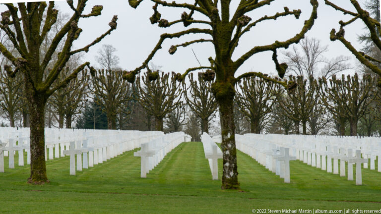 Long rows of white grave crosses recede in perspective between pollarded trees on bright green grass.