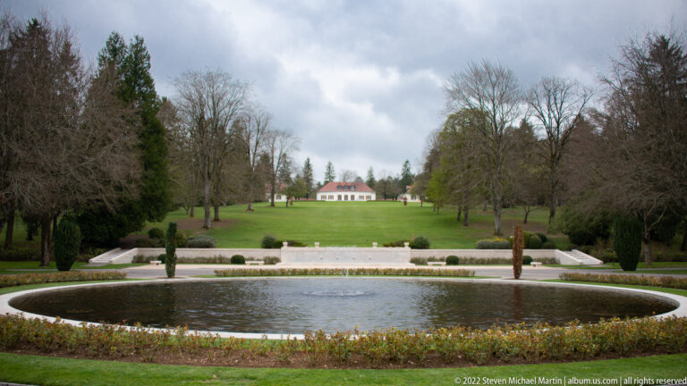 Large circular pool bordered by flowers and lawns, with a small building centered on the distant rise.