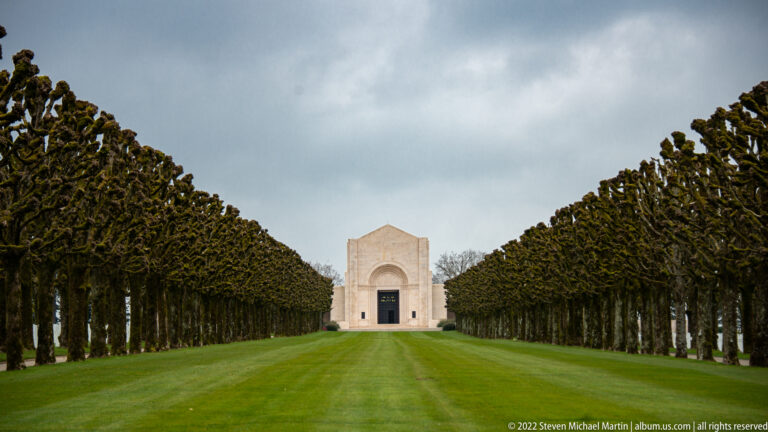 Pale stone cemetery chapel centered at the end of a long green lawn between pollarded tree rows.
