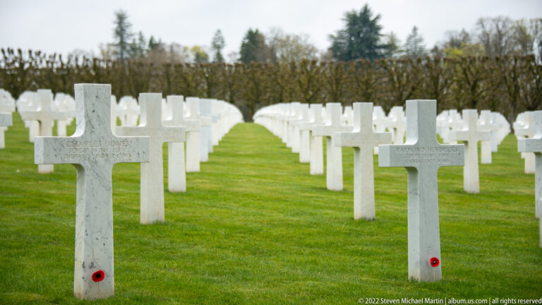 White marble crosses with small red remembrance poppies stand in rows across green grass.