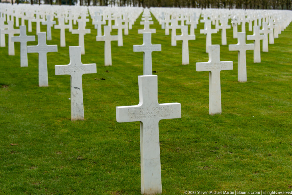 Single white marble cross in front of many aligned grave markers across a green cemetery lawn.