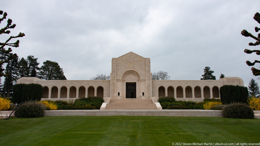 Front view of the pale stone cemetery chapel with a central gabled entrance and long side arcades.