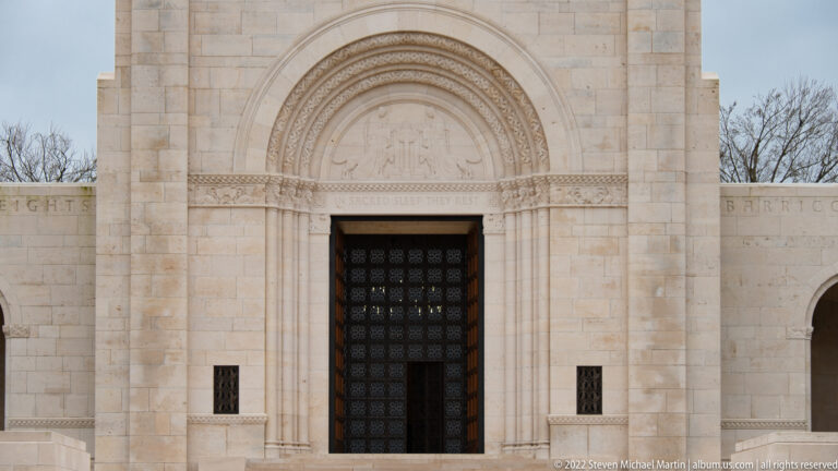 Ornate arched stone portal with carved bands, sculpted tympanum, and tall dark doors at the cemetery chapel.