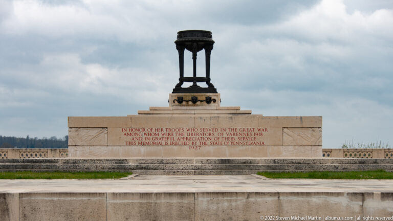 Low stone war memorial with carved dedication and a dark bronze openwork structure centered above.